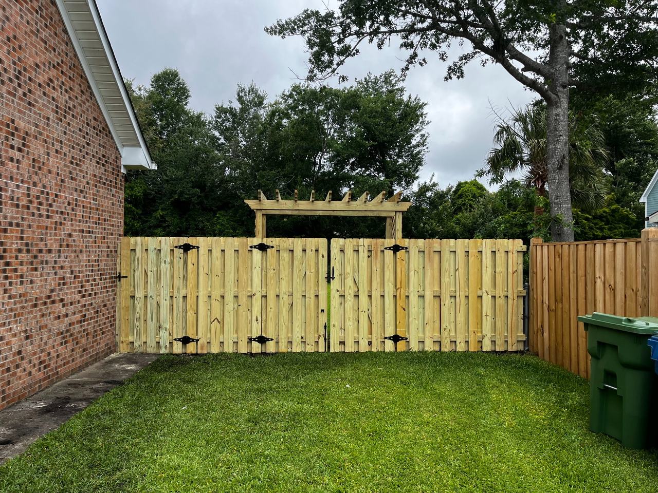 New wood privacy fence with double gates under a wooden pergola, next to a brick house