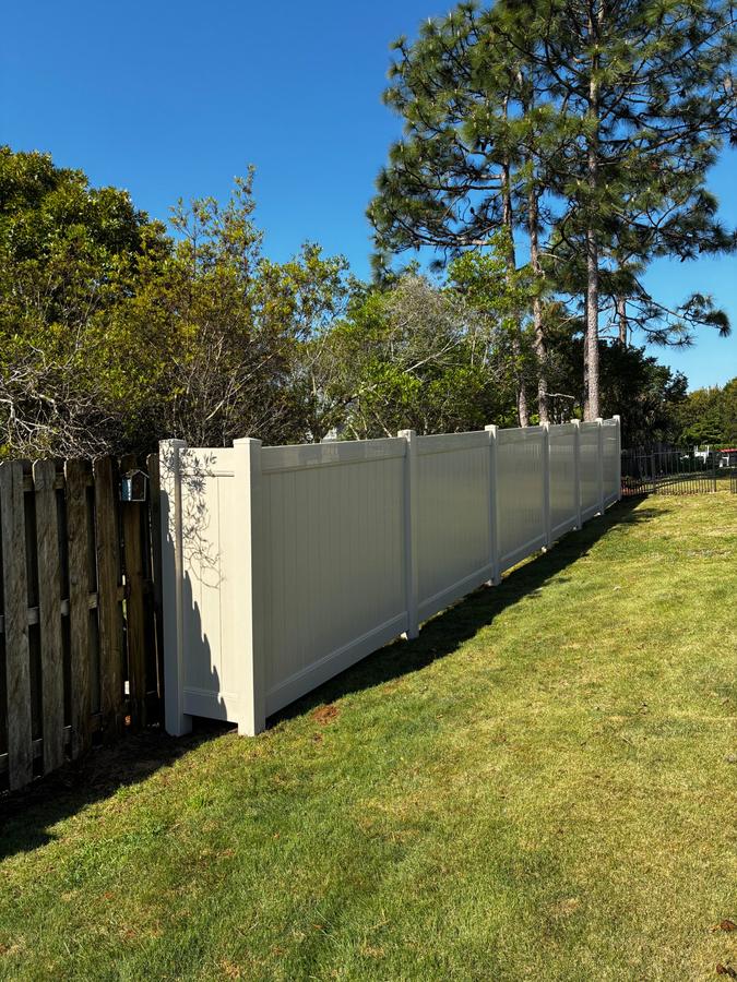 Tan vinyl privacy fence along a grassy yard under pine trees